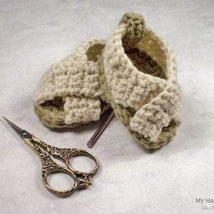 May include: A pair of crocheted baby sandals in beige and green with a criss-cross strap design. The sandals are lying on a white surface next to a pair of small scissors.
