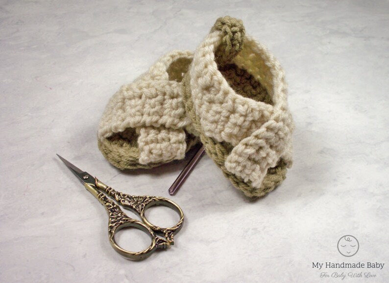 May include: A pair of crocheted baby sandals in beige and green with a criss-cross strap design. The sandals are lying on a white surface next to a pair of small scissors.