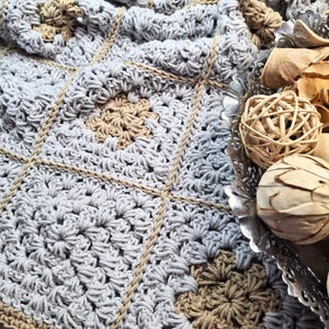 May include: A close-up of a handmade crochet blanket in shades of grey and tan. The blanket features a square pattern with floral-like designs in the centre of each square. The edges are outlined with a tan yarn, adding a decorative touch. The blanket is soft and textured.