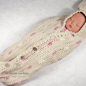 May include: A crocheted baby swaddle with pink and white stripes and a matching bow on the hood. The swaddle is made with a soft yarn and has buttons down the front. The baby is sleeping peacefully inside the swaddle.