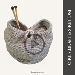 May include: A light gray crocheted yarn basket with a wooden ring closure. The basket contains yarn and two wooden knitting needles. The text "INCLUDES SUPPORT VIDEO" is on the right side of the image. The website bddpatterns.com is at the bottom.