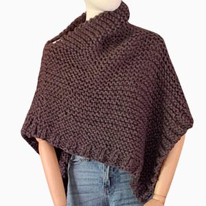 May include: A dark brown, chunky knit poncho with a high, ribbed turtleneck. The poncho has a loose, asymmetrical design, with a button closure on one shoulder. The garment is displayed on a mannequin, paired with blue jeans.