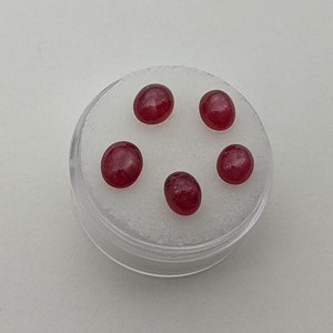 May include: Five round, red gemstones in a clear plastic container with a white background.