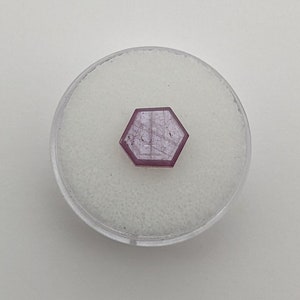 Könnte beinhalten: Ein sechseckiger, rosa Edelstein wird in einem transparenten, runden Behälter mit weißem, strukturiertem Innenraum präsentiert. Der Edelstein hat eine facettierte Oberfläche und ein durchscheinendes Aussehen. Der Behälter steht auf weißem Hintergrund.