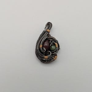 K&ouml;nnte beinhalten: Ein handgefertigter Anh&auml;nger mit Drahtwickeldesign. Der Anh&auml;nger hat einen dunklen Metallrahmen mit goldenen Akzenten, der einen weinroten Stein und einen kleinen gr&uuml;nen Stein umschlie&szlig;t. Die Gesamtform ist tropfenf&ouml;rmig.