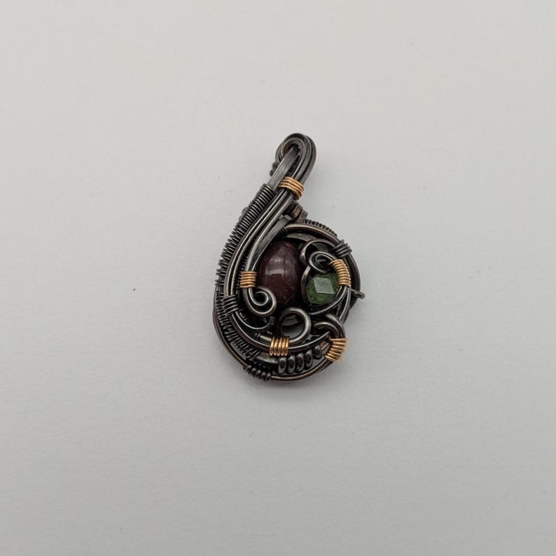 K&ouml;nnte beinhalten: Ein handgefertigter Anh&auml;nger mit Drahtwickeldesign. Der Anh&auml;nger hat einen dunklen Metallrahmen mit goldenen Akzenten, der einen weinroten Stein und einen kleinen gr&uuml;nen Stein umschlie&szlig;t. Die Gesamtform ist tropfenf&ouml;rmig.