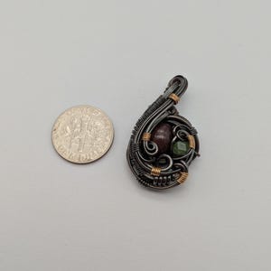 K&ouml;nnte beinhalten: Handgefertigter Anh&auml;nger mit einem Design aus dunklem Metalldraht. Der Anh&auml;nger hat einen weinroten Stein und einen gr&uuml;nen Stein, akzentuiert mit goldfarbenem Draht. Eine M&uuml;nze liegt neben dem Anh&auml;nger zum Gr&ouml;&szlig;envergleich.