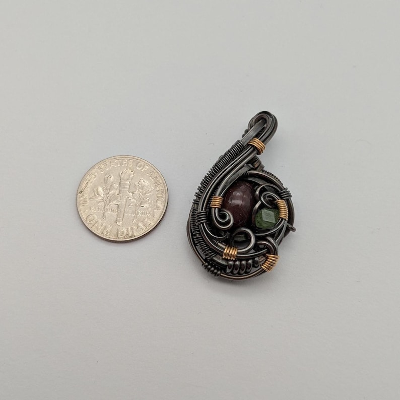 K&ouml;nnte beinhalten: Handgefertigter Anh&auml;nger mit einem Design aus dunklem Metalldraht. Der Anh&auml;nger hat einen weinroten Stein und einen gr&uuml;nen Stein, akzentuiert mit goldfarbenem Draht. Eine M&uuml;nze liegt neben dem Anh&auml;nger zum Gr&ouml;&szlig;envergleich.