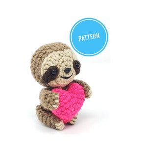 May include: A crocheted sloth holding a pink heart. The sloth is brown with black eyes and a smiling face. The heart is made of pink yarn.