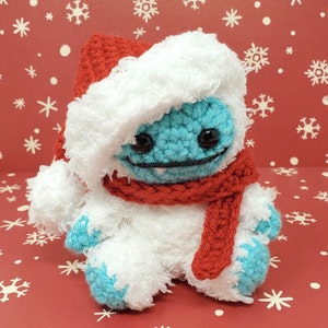 Op de afbeelding: Een handgemaakte gehaakte yeti-pop met een rode kerstmuts en sjaal. De yeti is voornamelijk wit met blauwe accenten op zijn gezicht en voeten. De achtergrond is rood met witte sneeuwvlokken.