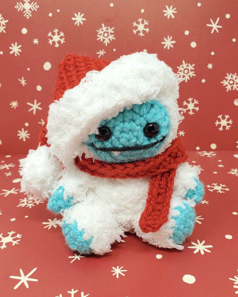 Op de afbeelding: Een handgemaakte gehaakte yeti-pop met een rode kerstmuts en sjaal. De yeti is voornamelijk wit met blauwe accenten op zijn gezicht en voeten. De achtergrond is rood met witte sneeuwvlokken.