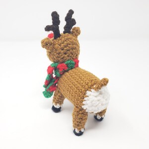 May include: A handmade crochet reindeer toy, viewed from the rear, with a brown body, black antlers, and a red nose. It wears a red and green scarf. The reindeer's rear is white, and its hooves are black.