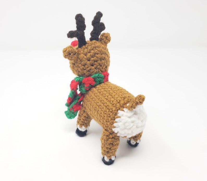 May include: A handmade crochet reindeer toy, viewed from the rear, with a brown body, black antlers, and a red nose. It wears a red and green scarf. The reindeer's rear is white, and its hooves are black.
