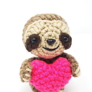 May include: A crocheted sloth holding a pink heart. The sloth has brown eyes and a smiling mouth.