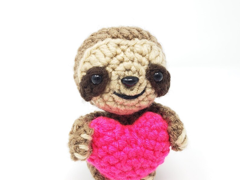 May include: A crocheted sloth holding a pink heart. The sloth has brown eyes and a smiling mouth.