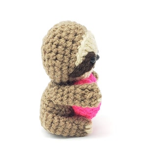 May include: A brown crocheted sloth holding a pink heart. The sloth is sitting on a white background.