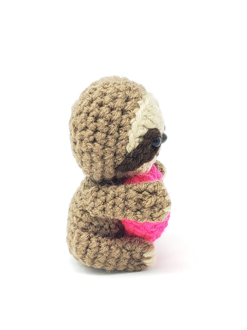 May include: A brown crocheted sloth holding a pink heart. The sloth is sitting on a white background.