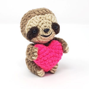 May include: A small crocheted sloth holding a bright pink heart. The sloth is brown and has a cute, smiling face.