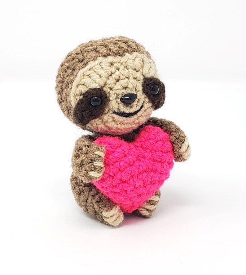May include: A small crocheted sloth holding a bright pink heart. The sloth is brown and has a cute, smiling face.