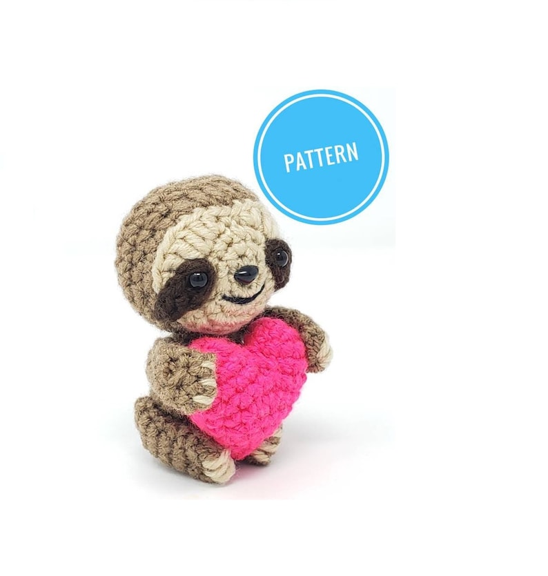 May include: A crocheted sloth holding a pink heart. The sloth is brown with black eyes and a smiling face. The heart is made of pink yarn.