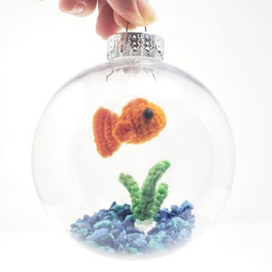 May include: A clear glass Christmas ornament with a crocheted orange fish, green seaweed, and blue gravel inside. The ornament is held by a silver cap and a hand. The fish has a black eye.