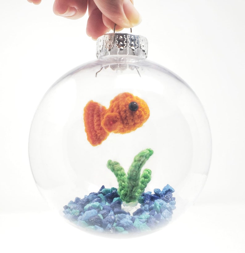 May include: A clear glass Christmas ornament with a crocheted orange fish, green seaweed, and blue gravel inside. The ornament is held by a silver cap and a hand. The fish has a black eye.