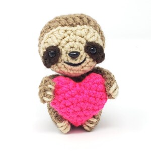 May include: A crocheted sloth holding a pink heart. The sloth has brown and beige fur, black eyes, and a smiling face.