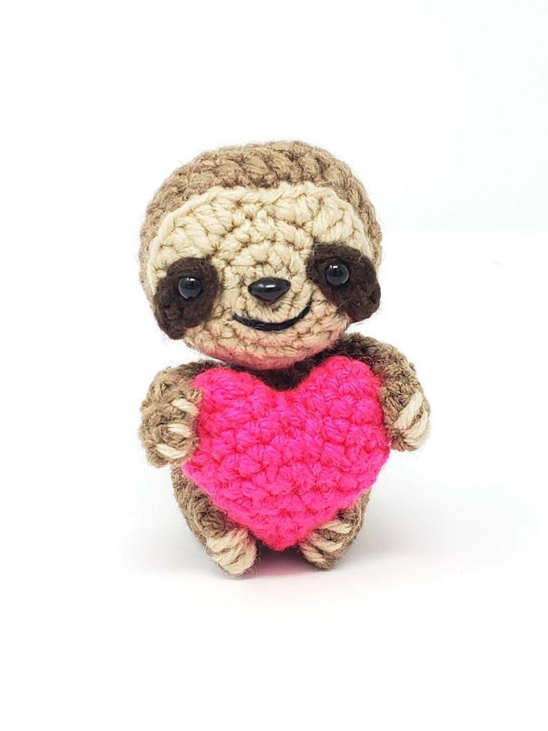 May include: A crocheted sloth holding a pink heart. The sloth has brown and beige fur, black eyes, and a smiling face.