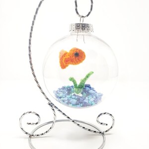 May include: A clear glass ornament with a crocheted orange goldfish, green plant, and blue gravel inside. The ornament hangs from a silver metal stand with decorative swirls. A unique, handmade decorative item.