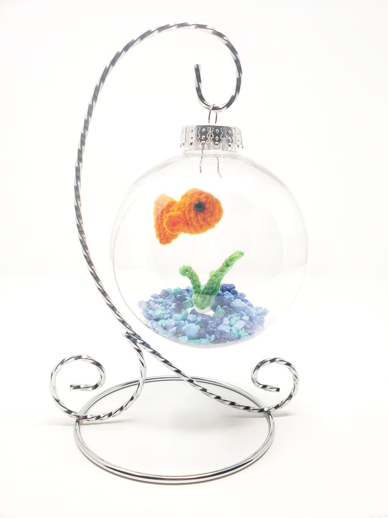 May include: A clear glass ornament with a crocheted orange goldfish, green plant, and blue gravel inside. The ornament hangs from a silver metal stand with decorative swirls. A unique, handmade decorative item.