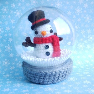 May include: A crocheted snowman inside a clear snow globe. The snowman wears a black top hat, a red scarf, and has black button eyes. The base of the globe is gray, and the background is light blue with white snowflakes.