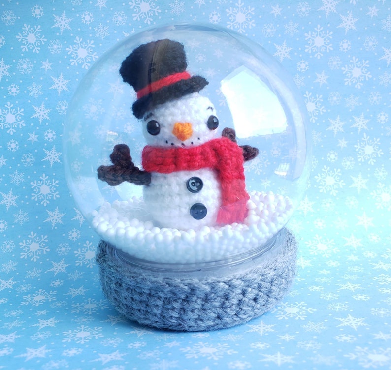 May include: A crocheted snowman inside a clear snow globe. The snowman wears a black top hat, a red scarf, and has black button eyes. The base of the globe is gray, and the background is light blue with white snowflakes.