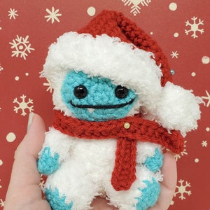 Op de afbeelding: Handgemaakte gehaakte yeti-pop met een rode kerstmuts en sjaal. De yeti is lichtblauw met zwarte ogen en een gestikte glimlach. De hoed en sjaal zijn rood, en het lichaam is wit. De achtergrond is rood met witte sneeuwvlokken.