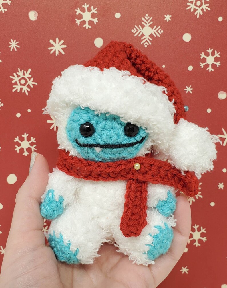 Op de afbeelding: Handgemaakte gehaakte yeti-pop met een rode kerstmuts en sjaal. De yeti is lichtblauw met zwarte ogen en een gestikte glimlach. De hoed en sjaal zijn rood, en het lichaam is wit. De achtergrond is rood met witte sneeuwvlokken.