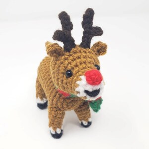 May include: A handmade crochet reindeer with a brown body, dark brown antlers, and a red nose. The reindeer has a white muzzle, black eyes, and a green and red scarf. The reindeer is standing on a white surface.