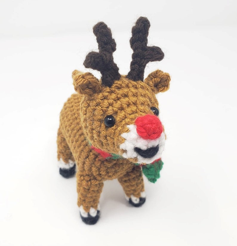 May include: A handmade crochet reindeer with a brown body, dark brown antlers, and a red nose. The reindeer has a white muzzle, black eyes, and a green and red scarf. The reindeer is standing on a white surface.