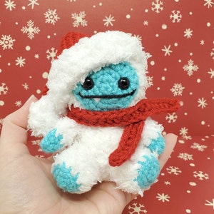 Op de afbeelding: Een handgemaakte gehaakte sneeuwman met een rode kerstmuts en sjaal. De sneeuwman heeft een blauw gezicht en voeten, zwarte ogen en een gestikte glimlach. De sneeuwman is ongeveer 10 cm hoog.