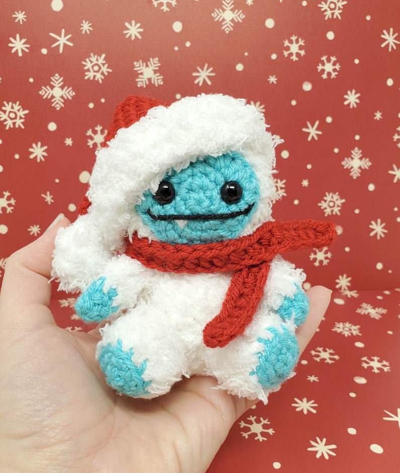 Op de afbeelding: Een handgemaakte gehaakte sneeuwman met een rode kerstmuts en sjaal. De sneeuwman heeft een blauw gezicht en voeten, zwarte ogen en een gestikte glimlach. De sneeuwman is ongeveer 10 cm hoog.