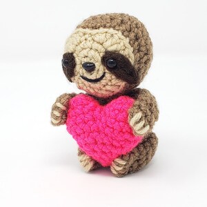 May include: A crocheted sloth holding a pink heart. The sloth has brown fur, black eyes, and a smiling face.