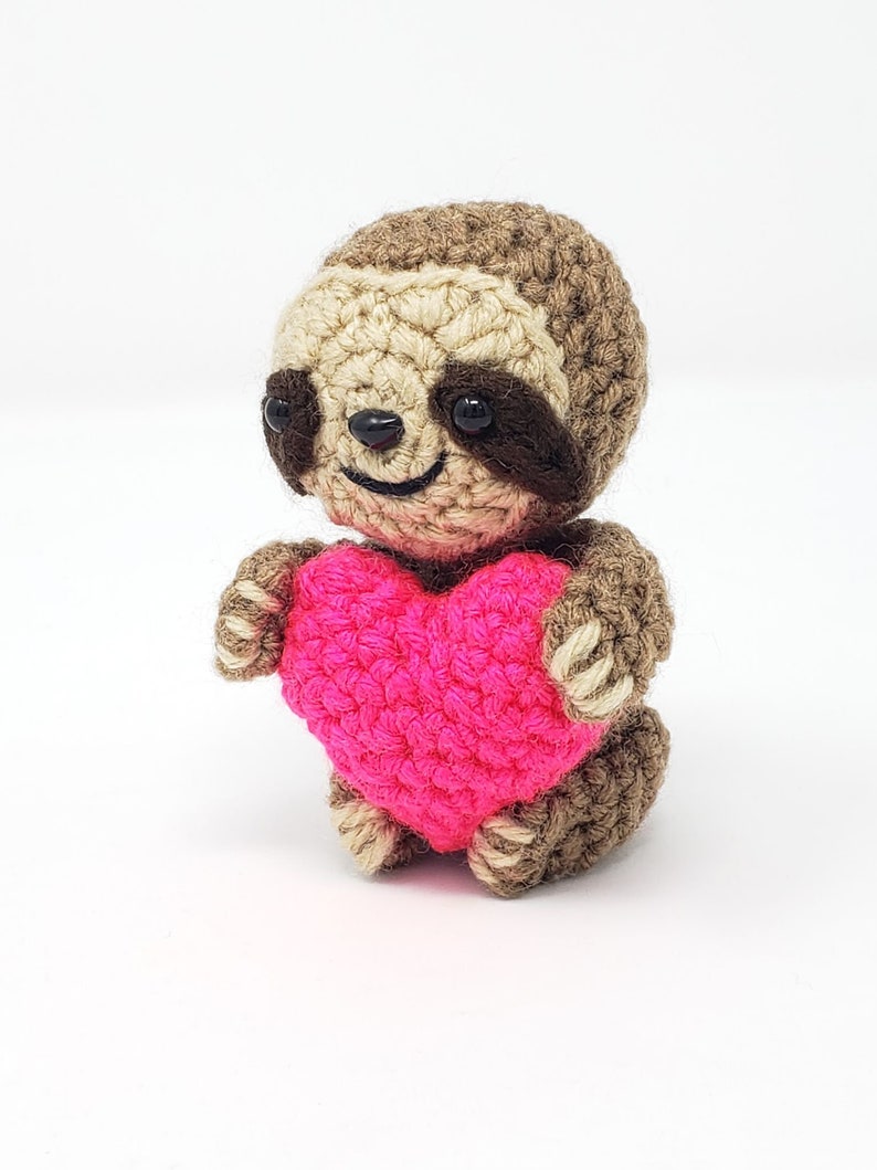 May include: A crocheted sloth holding a pink heart. The sloth has brown fur, black eyes, and a smiling face.