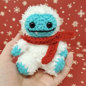 Op de afbeelding: Een handgemaakte gehaakte yeti-pop met een witte body, turquoise gezicht en voeten en een rode sjaal. De pop heeft zwarte ogen en een lachende mond. De achtergrond is een rode ondergrond met witte sneeuwvlokken.