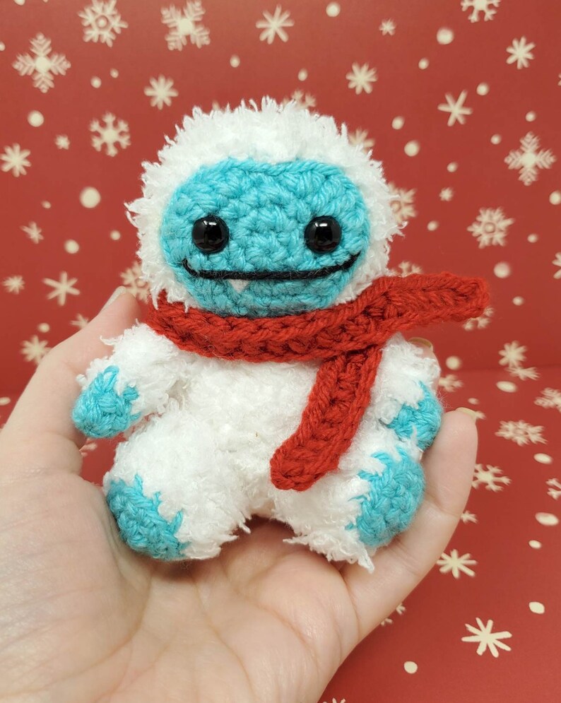 Op de afbeelding: Een handgemaakte gehaakte yeti-pop met een witte body, turquoise gezicht en voeten en een rode sjaal. De pop heeft zwarte ogen en een lachende mond. De achtergrond is een rode ondergrond met witte sneeuwvlokken.