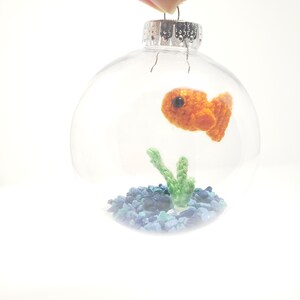 May include: Clear glass Christmas ornament with a crocheted orange goldfish, green plant, and blue and green gravel inside. The ornament is held by a hand against a white background. The ornament has a silver metal cap.