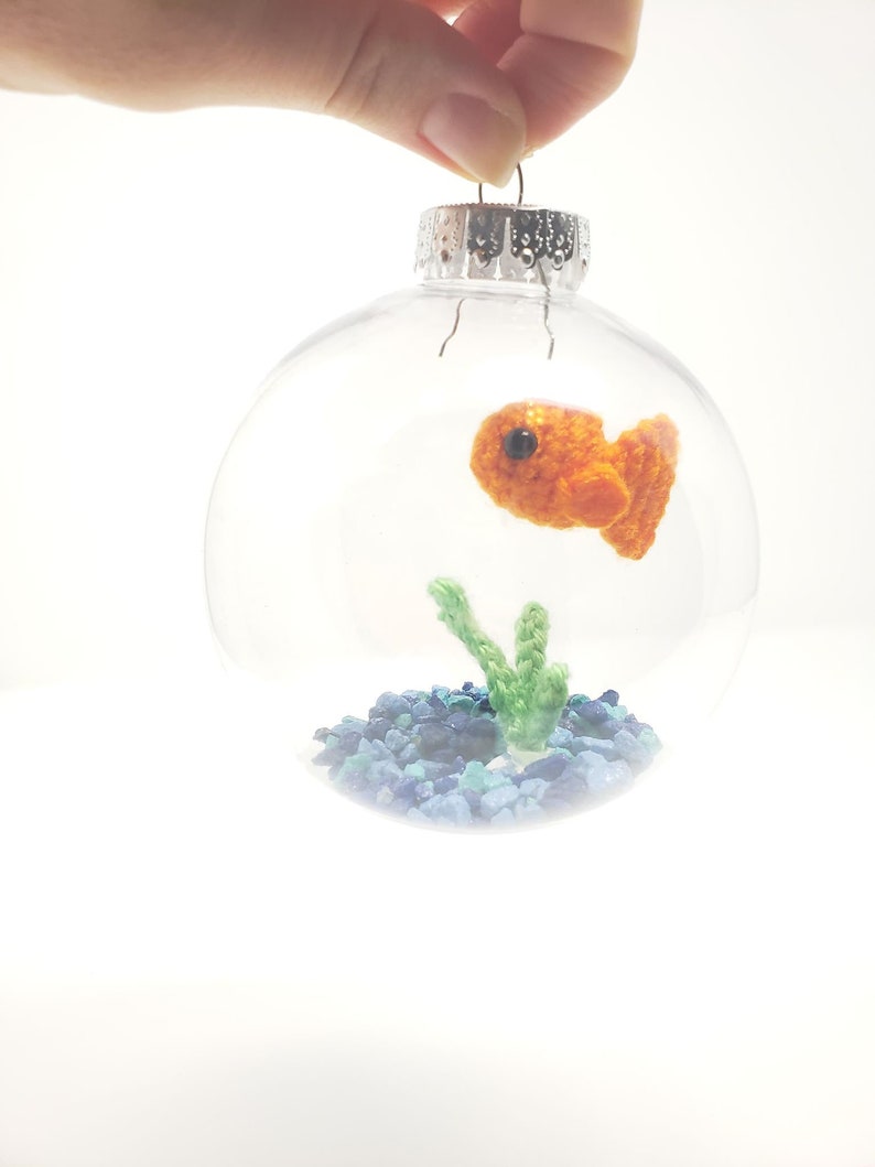 May include: Clear glass Christmas ornament with a crocheted orange goldfish, green plant, and blue and green gravel inside. The ornament is held by a hand against a white background. The ornament has a silver metal cap.