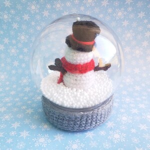 May include: A clear snow globe featuring a crocheted snowman with a red scarf, black hat, and twig arms. The snowman sits on a bed of white foam inside the globe, which is set on a gray crocheted base. The background is light blue with white snowflakes.