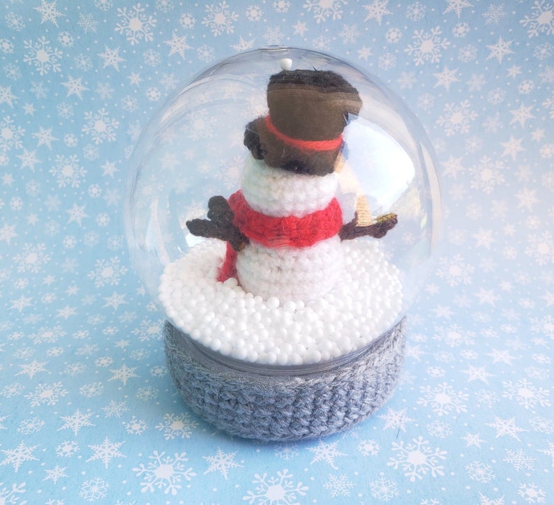 May include: A clear snow globe featuring a crocheted snowman with a red scarf, black hat, and twig arms. The snowman sits on a bed of white foam inside the globe, which is set on a gray crocheted base. The background is light blue with white snowflakes.