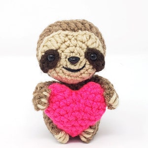May include: A small crocheted sloth holding a pink heart. The sloth is brown with black eyes and a smiling mouth.