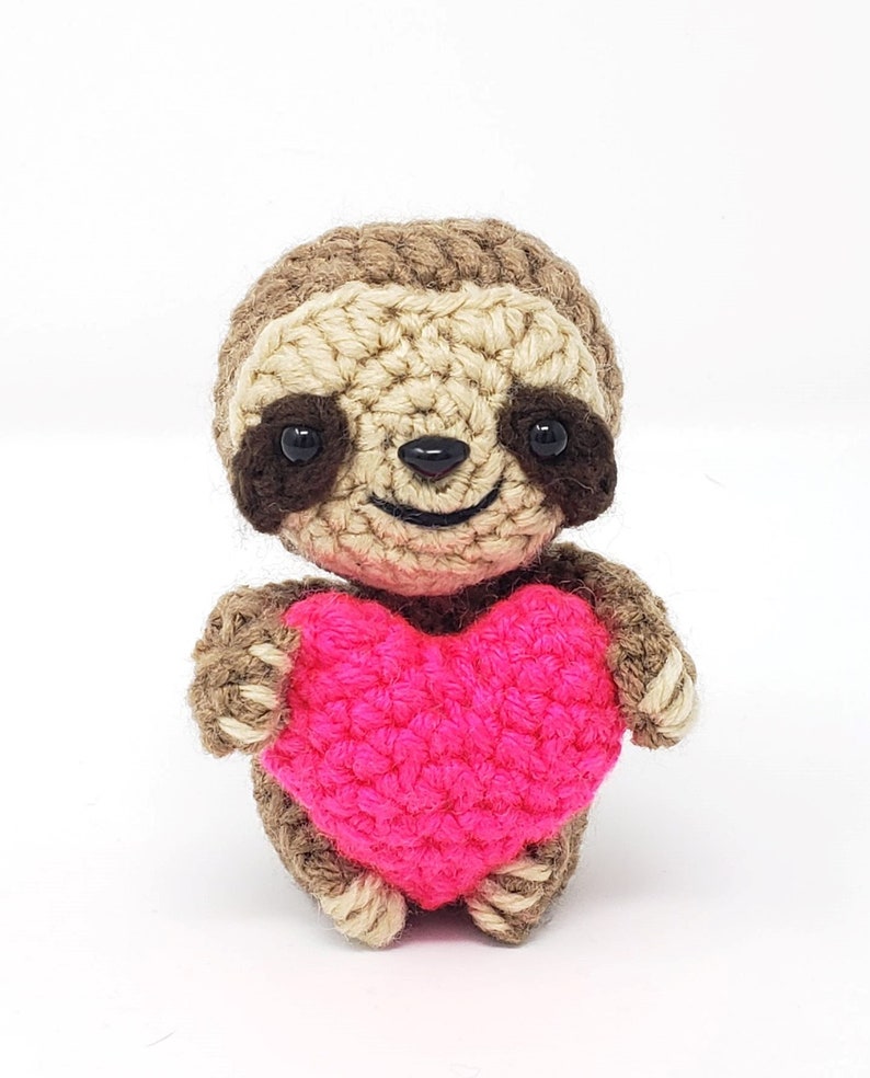 May include: A small crocheted sloth holding a pink heart. The sloth is brown with black eyes and a smiling mouth.