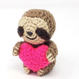 May include: A crocheted sloth holding a pink heart. The sloth is brown and has black eyes.