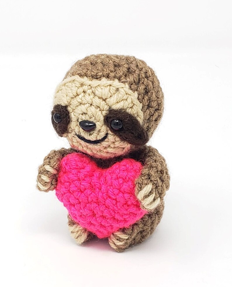 May include: A crocheted sloth holding a pink heart. The sloth is brown and has black eyes.
